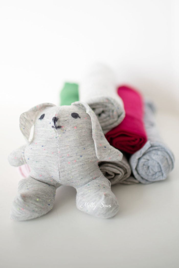 Gray bunny stuffy sewn from knit scrap fabric sitting near rolls of knit fabric scraps