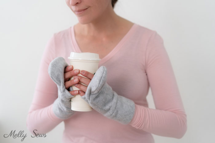 Woman in a pink shirt holding a coffee cup while wearing gray fleece mittens with fingerless flaps