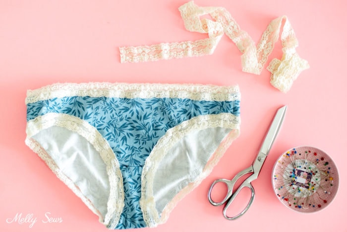 Blue floral women's underwear with ivory lace elastic trim on a pink background with scissors and a pin dish