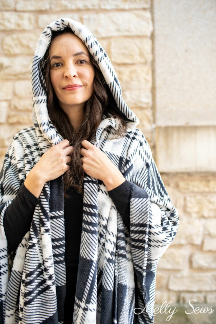 Woman wearing a black and white hooded blanket she made in front of a limestone walls