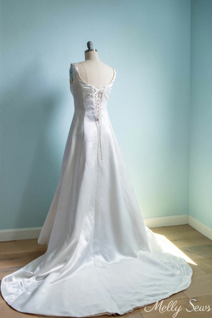 Back view of a white satin gown with a train and a lace up back on a dress form in front of a pale aqua wall