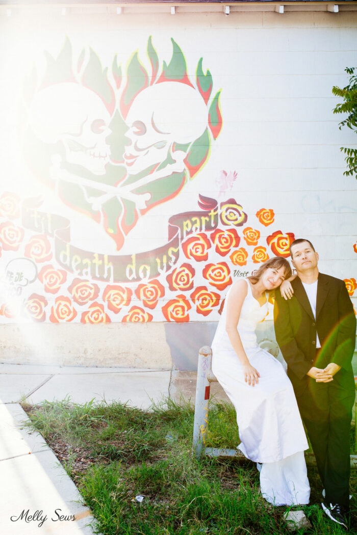 Woman in a wedding dress leaning on man in a tuxedo jacket in front of a mural with skulls in a heart and the words Til Death Do Us Part
