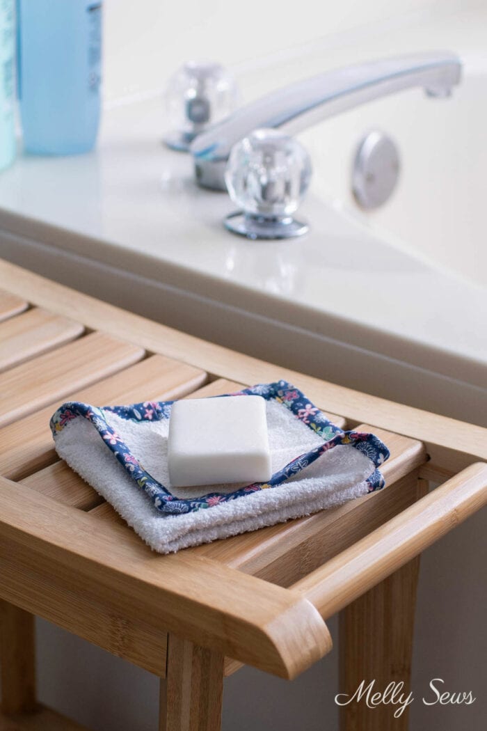 A washcloth with navy floral trim on a wooden bench with a bar of soap next to a tub