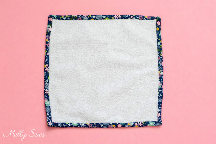A square white towel wash cloth with navy blue floral trim on a pink background