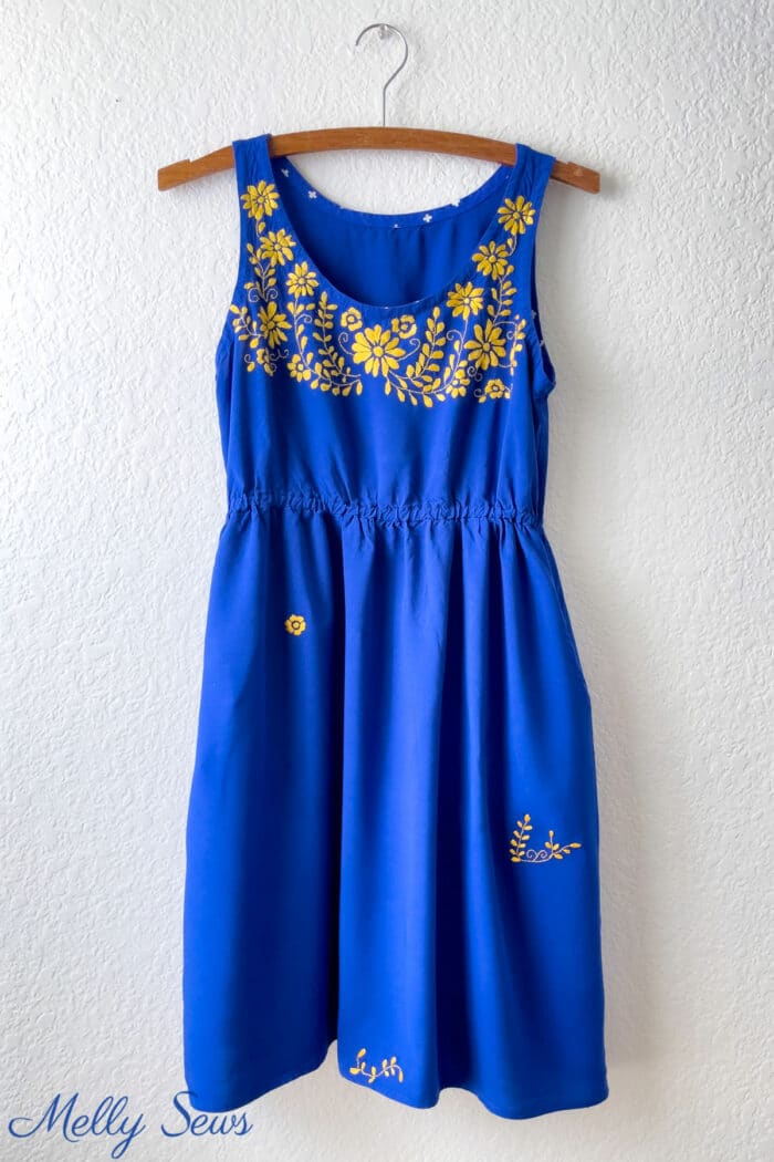 A blue dress with gold embroidery hangs on a wall