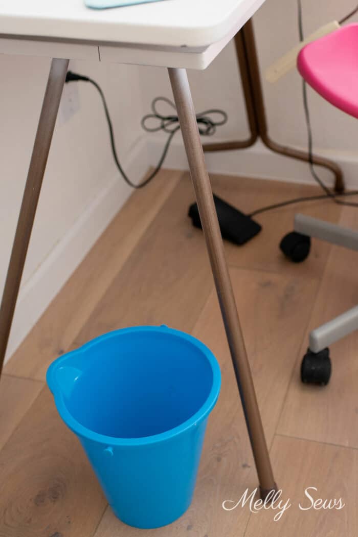 Foot pedal of a sewing machine under a table next to a blue thread bucket