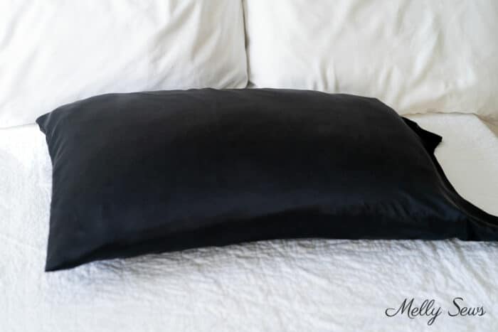 Black silk pillowcase on a pillow resting on a white bedspread with white cotton pillowcases in the background