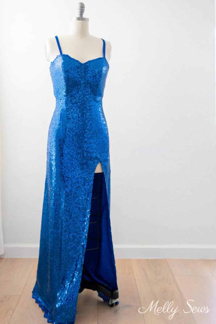 Blue floor length sequin prom dress with a v neckline, spaghetti straps and leg slit displayed on a dress form
